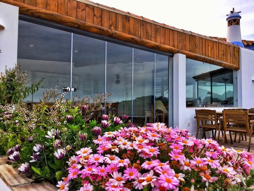 A modern building with large glass windows, wooden deck, and a variety of flowers in the foreground.