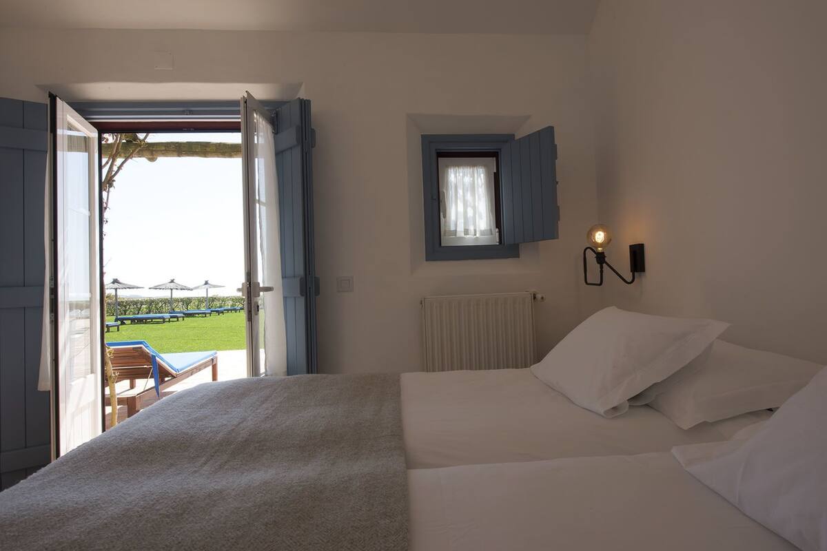 A bedroom with a bed, a window with blue shutters, a wall-mounted light, and a view of a garden outside.
