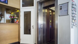 A hotel reception area with a reception desk, a wall-mounted information board, and an elevator.