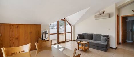 A spacious living area with a high ceiling, a dining table with chairs, a gray sofa, and a television on a stand.
