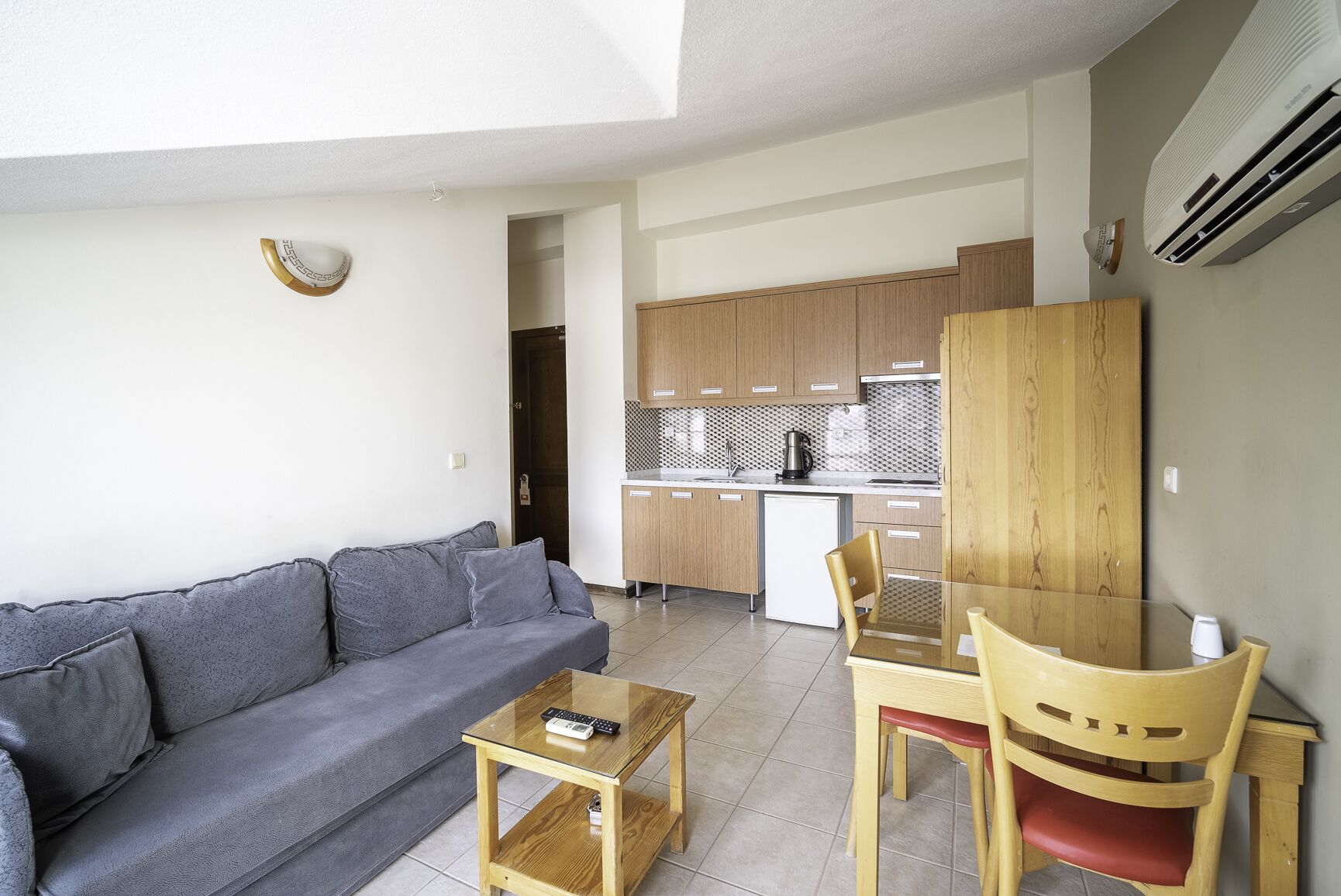 A modern living room with a grey sofa, a wooden coffee table, and a glass dining table with two chairs. A kitchen with wooden cabinets and a white refrigerator is visible in the background.