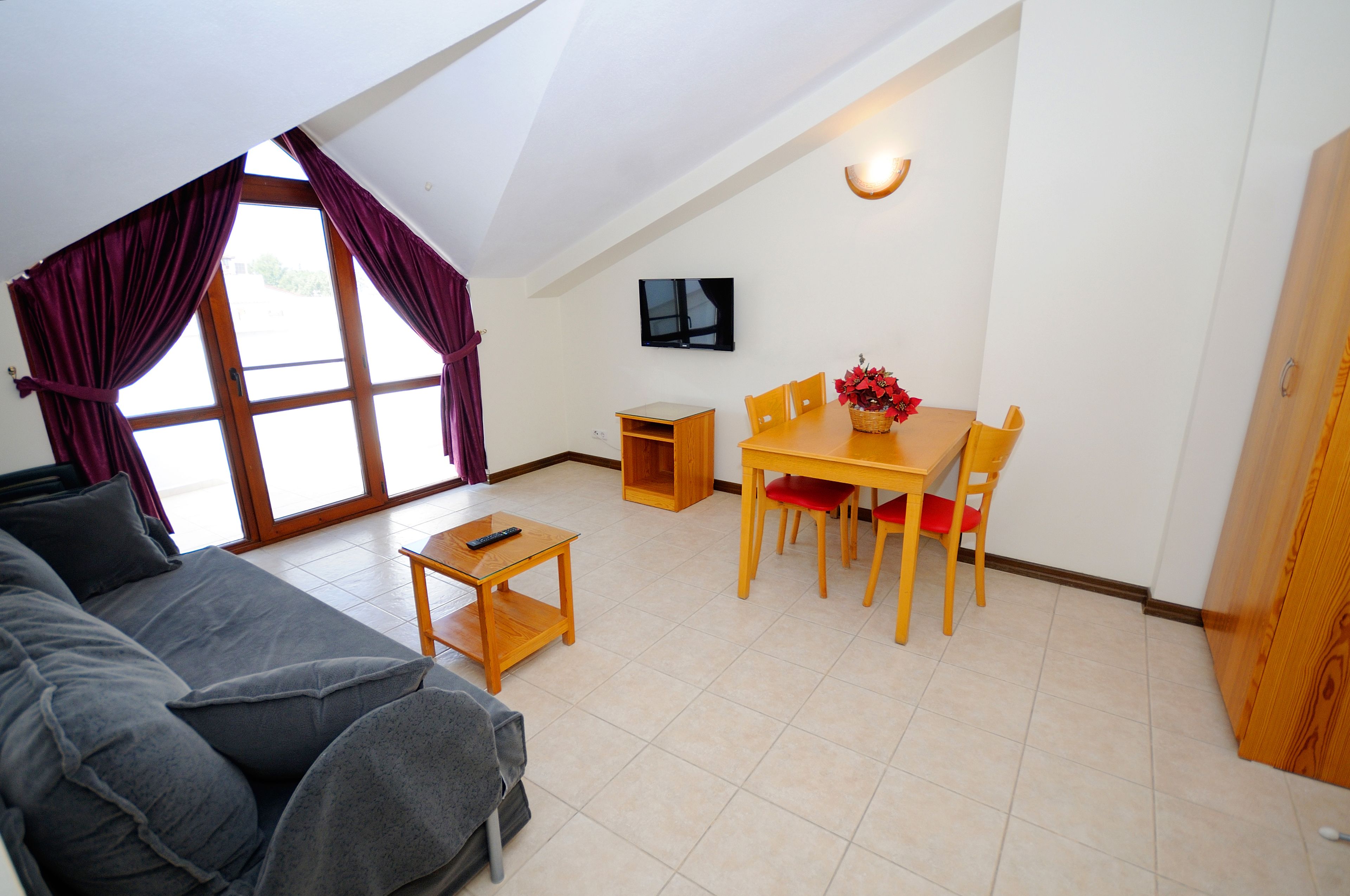 A cozy living room with a sofa, a dining table with chairs, a TV mounted on the wall, and a window with curtains.
