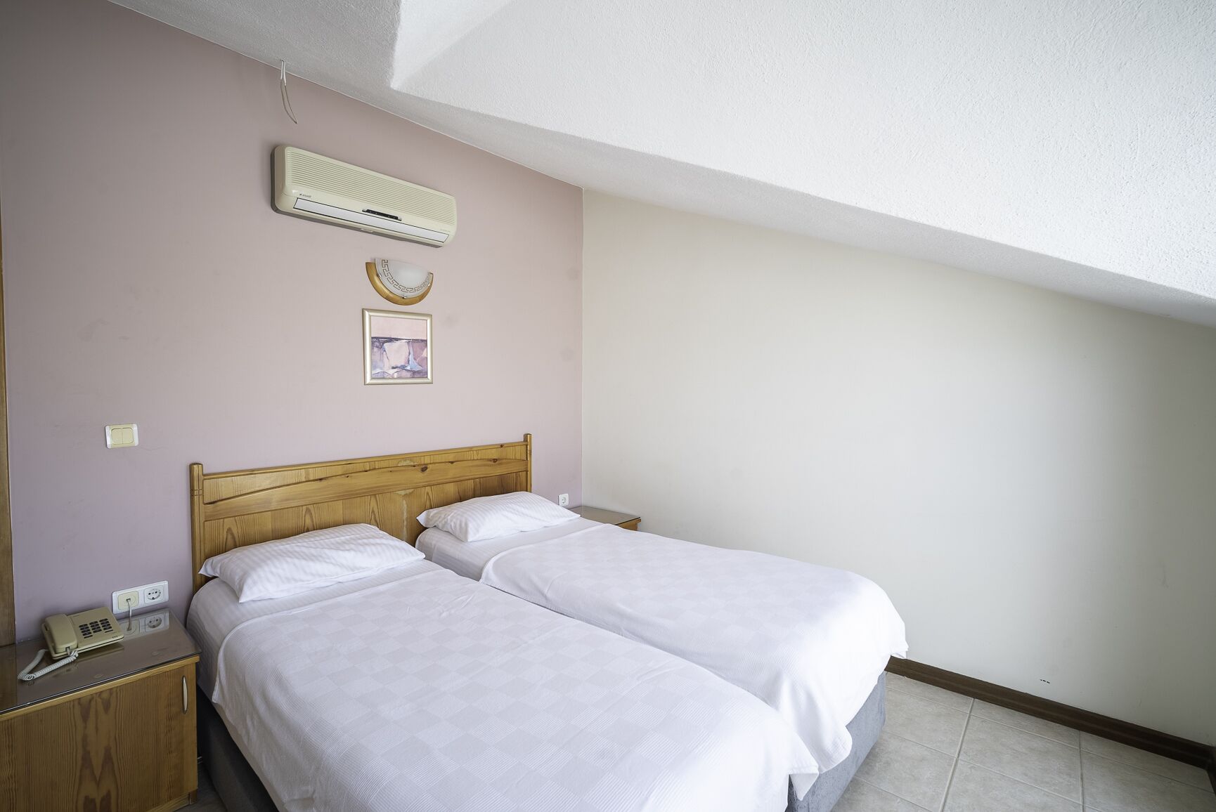 A bedroom with two single beds, a wooden headboard, a bedside table with a telephone, a wall-mounted air conditioner, and a framed picture.