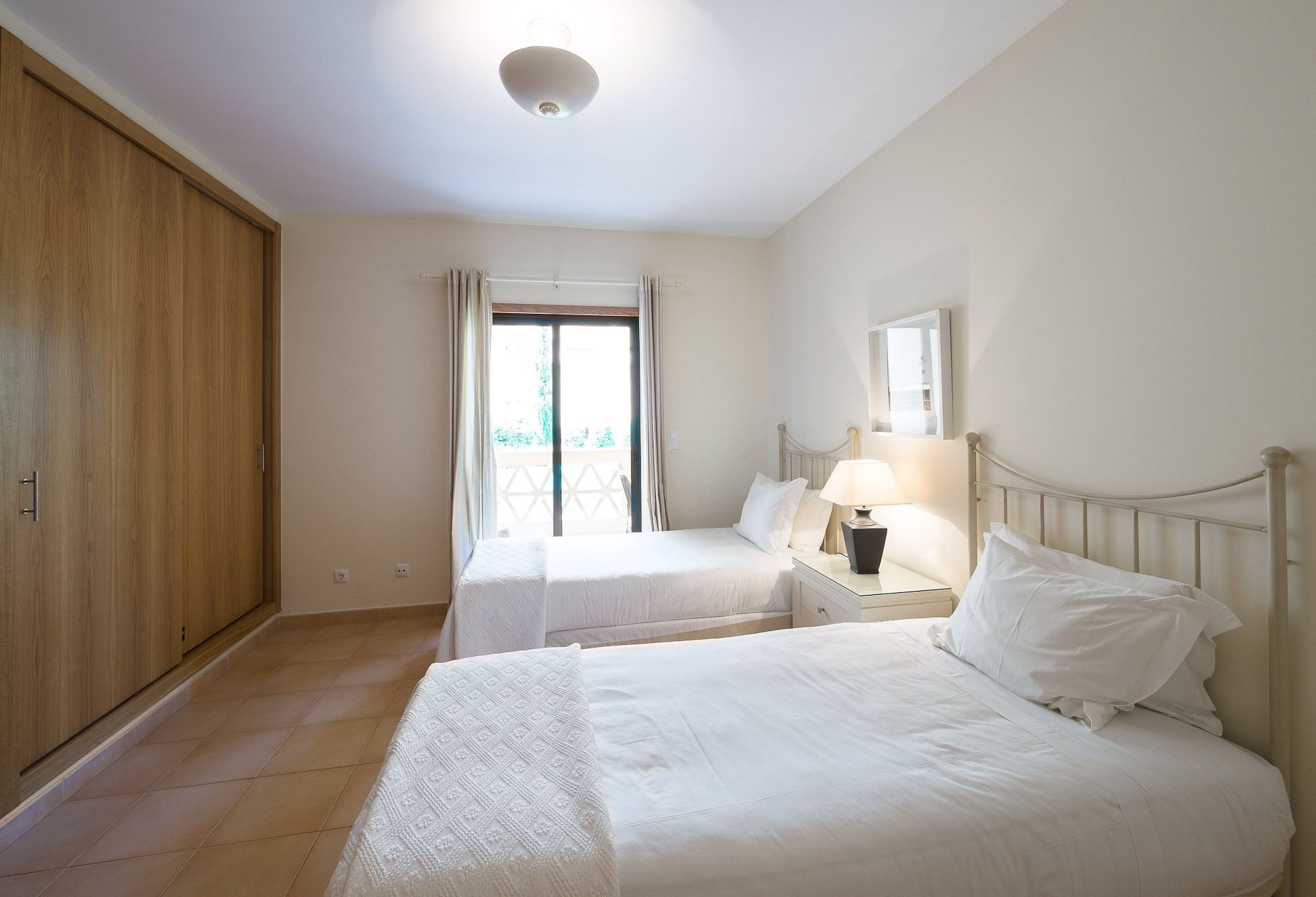 A hotel room with two beds, a wooden wardrobe, a window with curtains, a bedside table with a lamp, and a ceiling light.