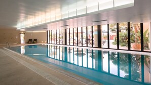 Piscina interior com paredes de vidro transparente, piso em azulejo e vista para área externa com assentos e vegetação.
