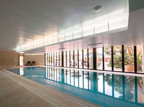 Piscina interior com paredes de vidro transparente, piso em azulejo e vista para área externa com assentos e vegetação.