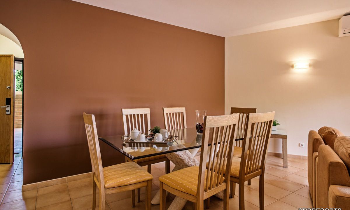 A dining area with a glass table and wooden chairs, a sofa, and a wall-mounted light fixture.