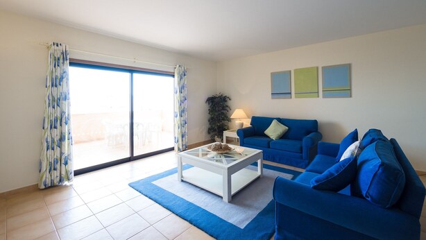 Sala de estar com um sofá azul, uma mesa de centro branca e uma janela grande com cortinas.