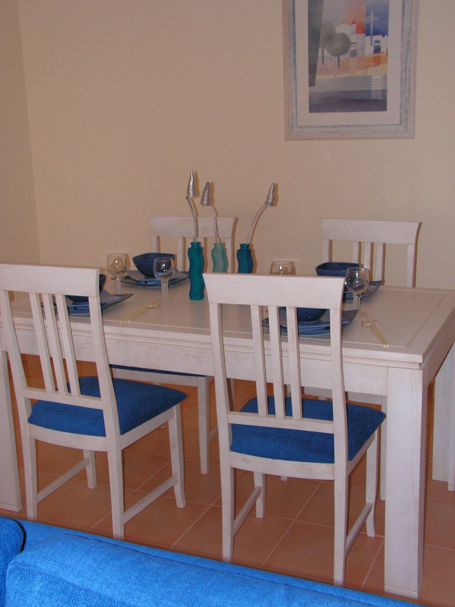 A dining area with a white table and chairs, blue cushions, and a painting on the wall.