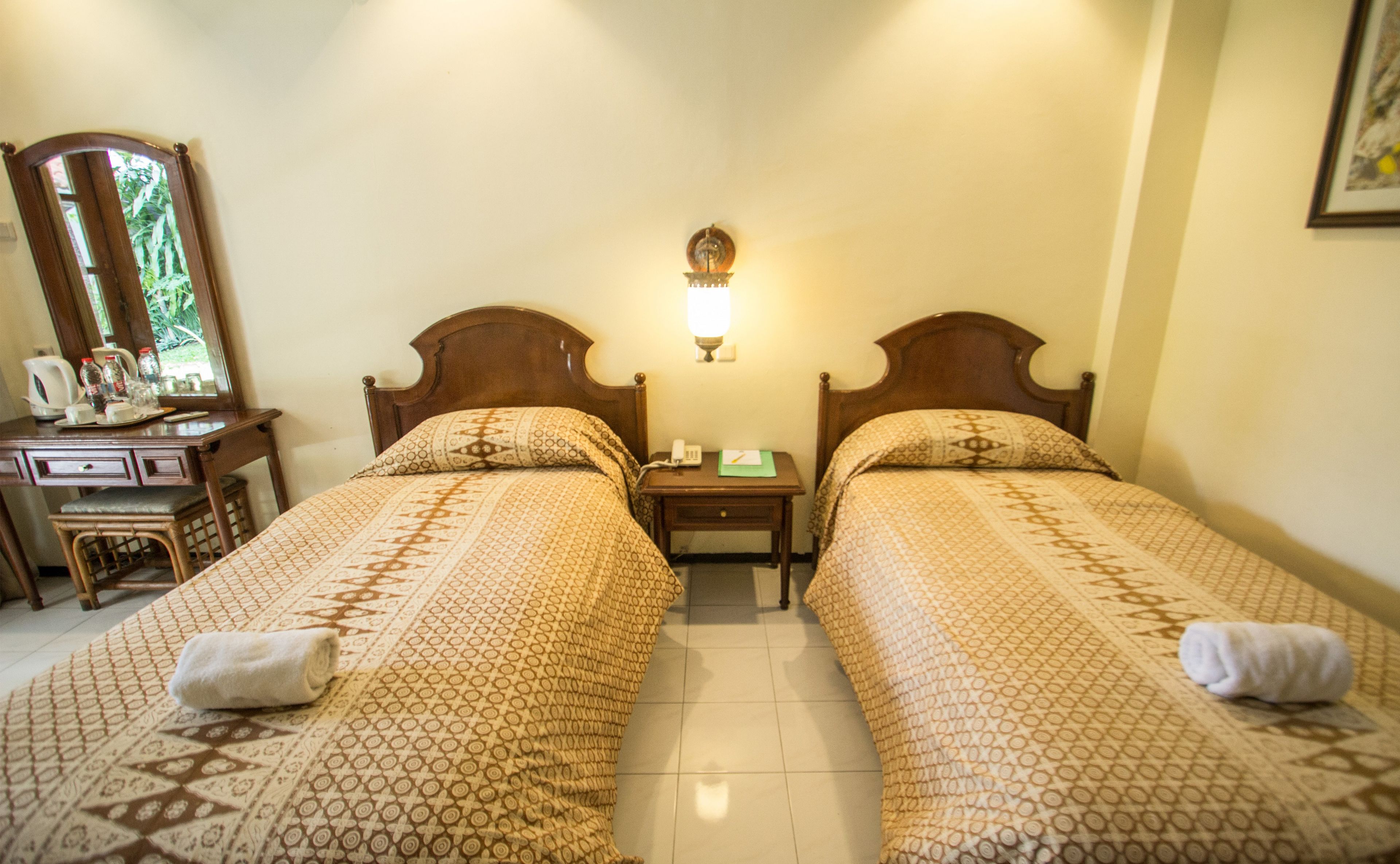 Habitación de hotel con dos camas, una mesita de noche con lámpara, un espejo, un escritorio con silla y una ventana que da a zonas verdes.