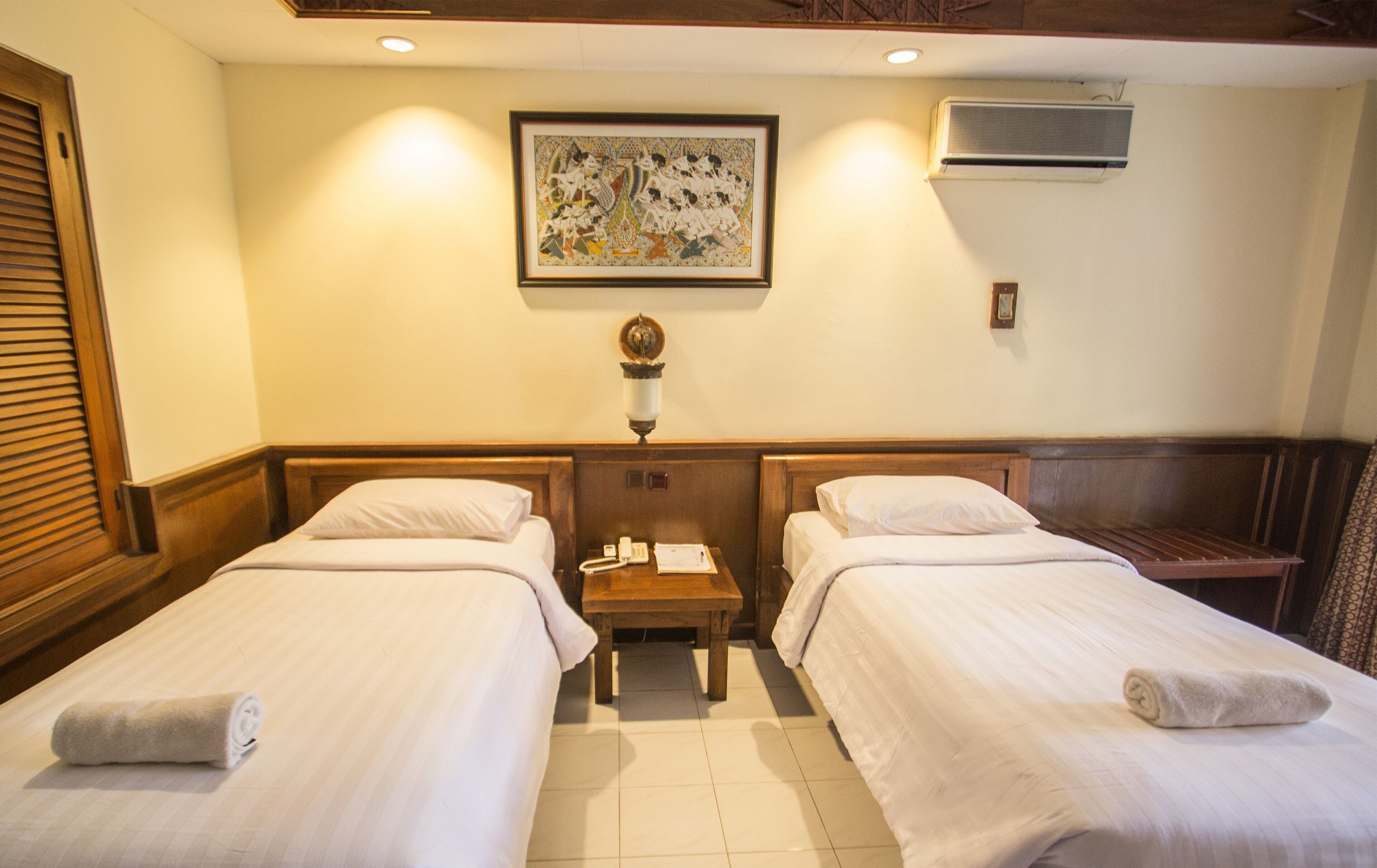 A hotel room with two single beds, a small wooden nightstand, a framed artwork on the wall, and an air conditioning unit.