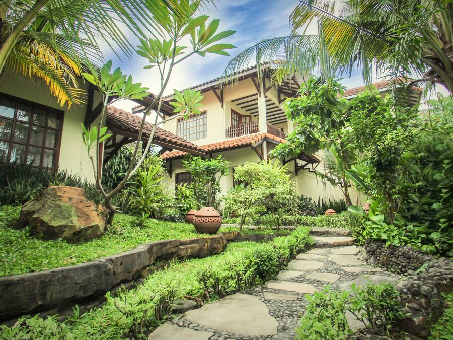 A tropical garden with a stone pathway leading to a building with a tiled roof and large windows.