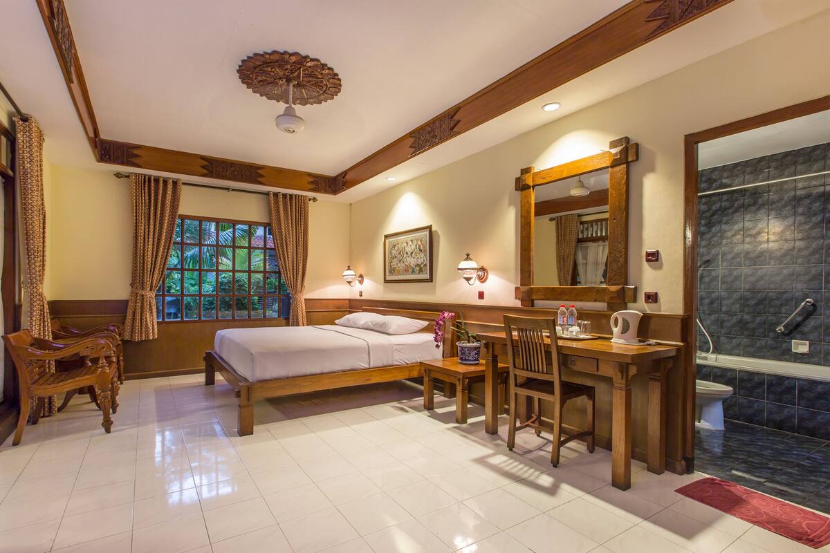 Habitación de hotel con cama, muebles de madera, escritorio, baño con ducha y ventana con cortinas.