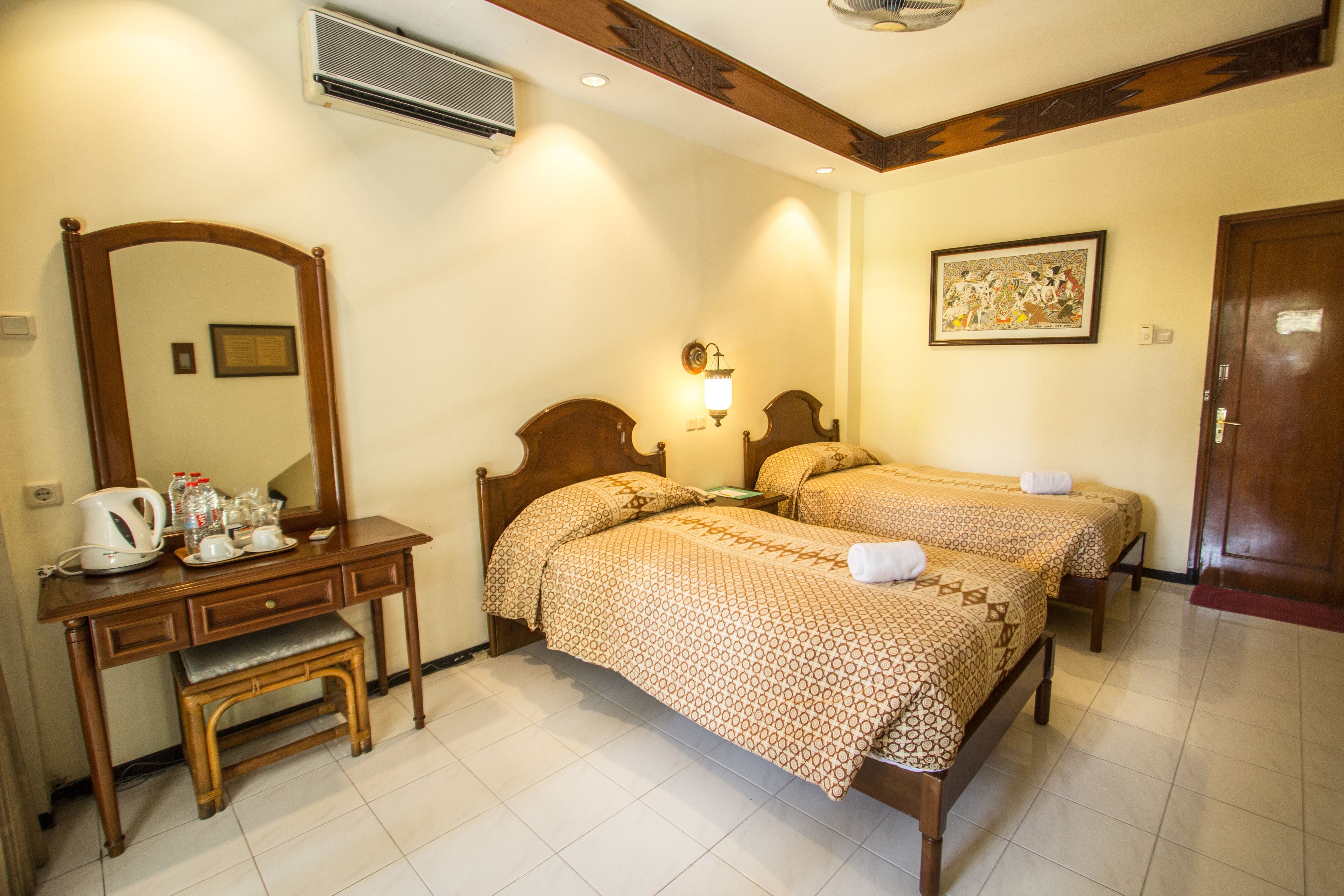A hotel room with two wooden beds, a wooden desk with a bench, a mirror, a wall-mounted air conditioner, and a framed picture on the wall.