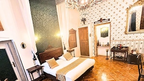 A room with a large bed, a chandelier, patterned wallpaper, and a wooden floor.