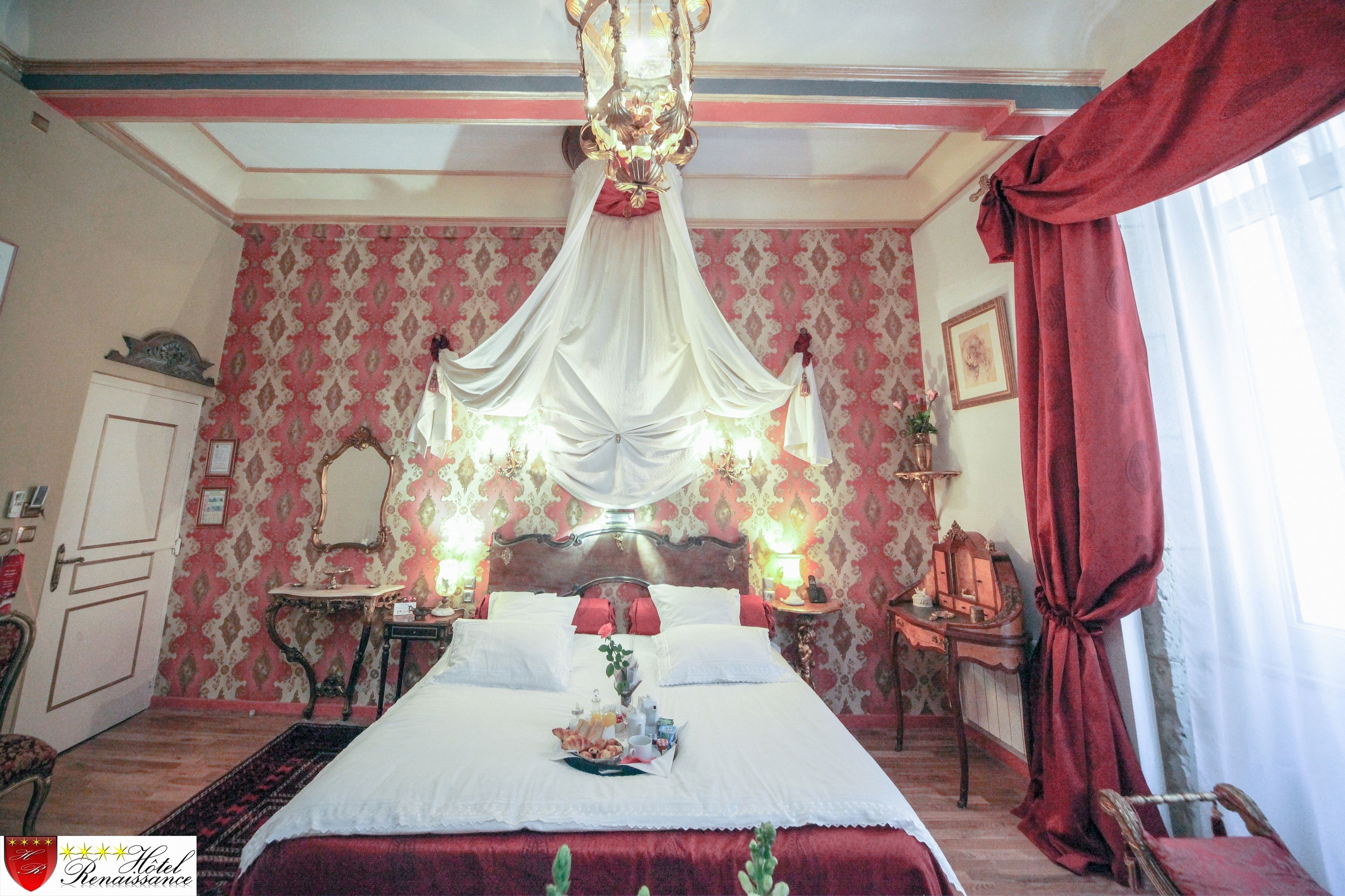 Habitación de hotel con una cama grande, un candelabro, un espejo, una mesita con una lámpara y una cortina roja.