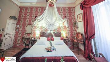 Habitación de hotel con una cama grande, un candelabro, un espejo, una mesita con una lámpara y una cortina roja.