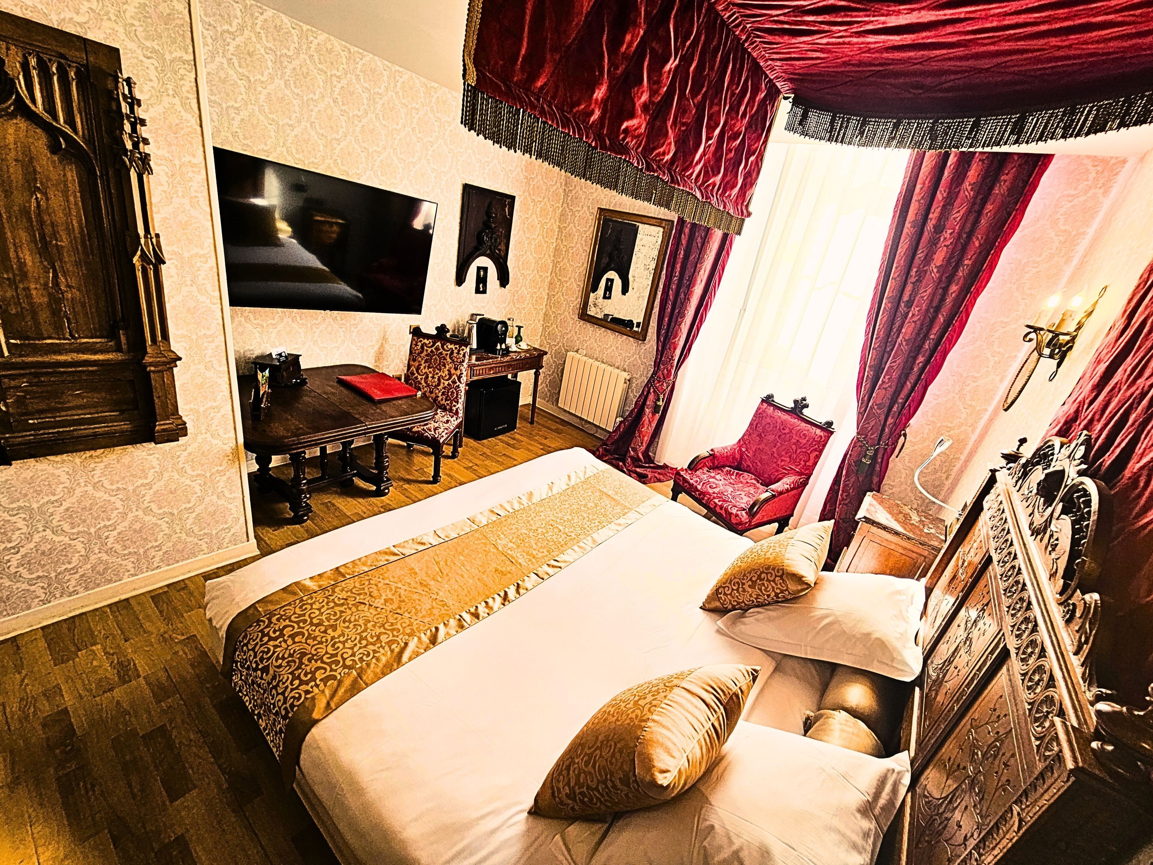 A room with a large bed, a wooden dresser, a red armchair, a desk with a chair, and a television mounted on the wall.
