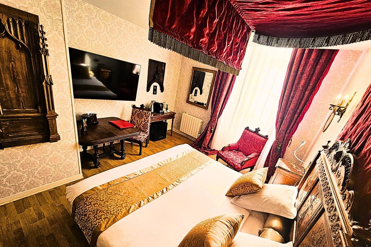 A room with a large bed, a wooden dresser, a red armchair, a desk with a chair, and a television mounted on the wall.