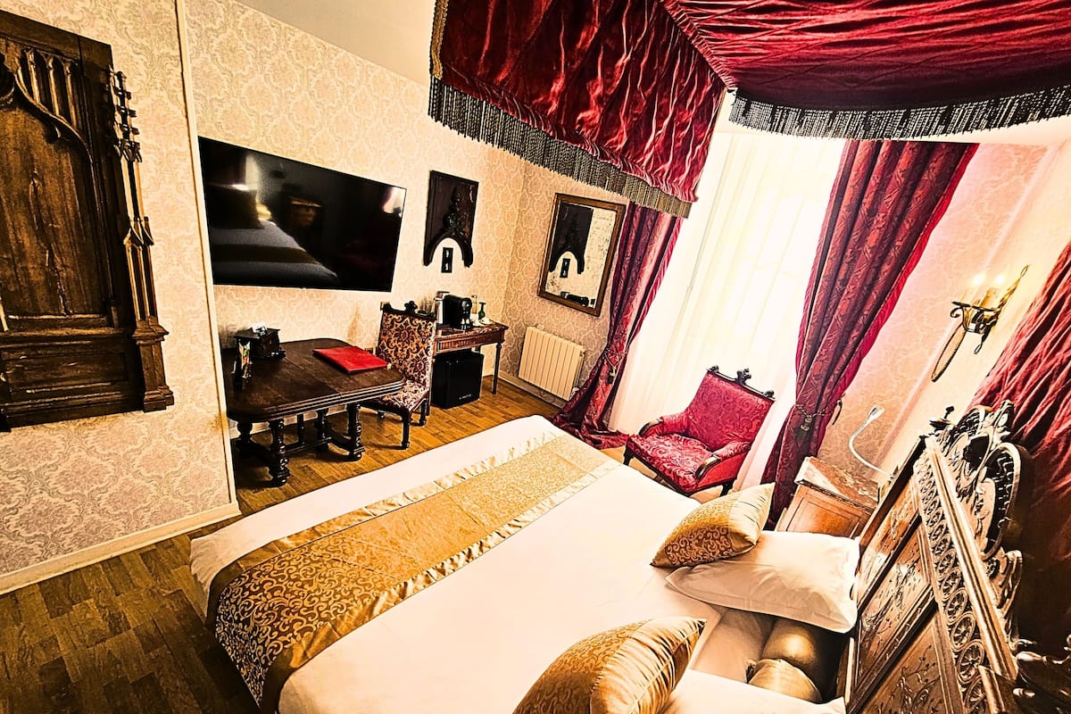 A room with a large bed, a wooden dresser, a red armchair, a desk with a chair, and a television mounted on the wall.