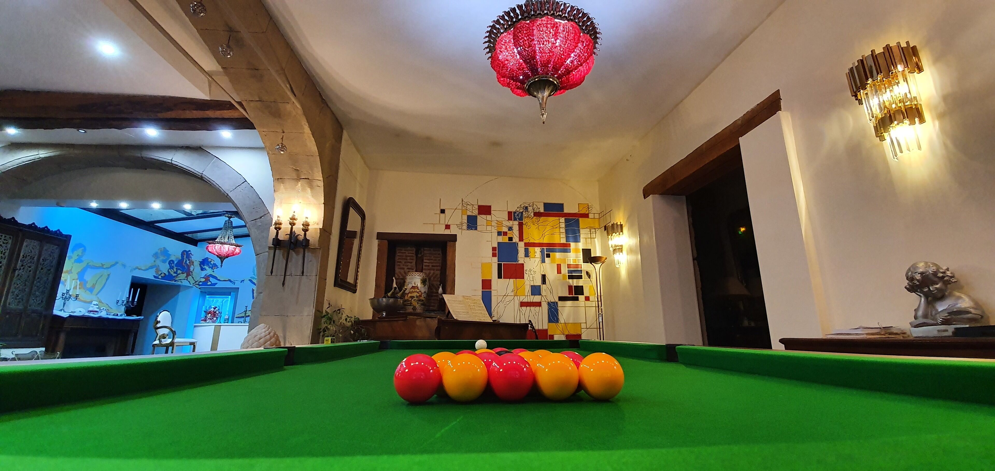 Una mesa de billar con bolas en una habitación con techo decorativo, una pared con obras de arte coloridas y una estatua.