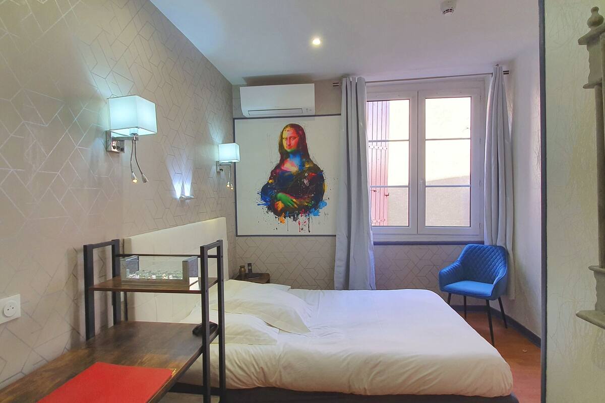A hotel room with a bed, a desk with a chair, a window with curtains, and a wall-mounted air conditioner.