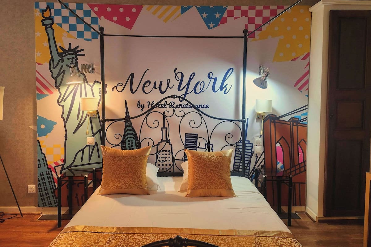 A hotel room with a bed, two decorative pillows, a wall mural of New York City landmarks, and a black metal headboard.