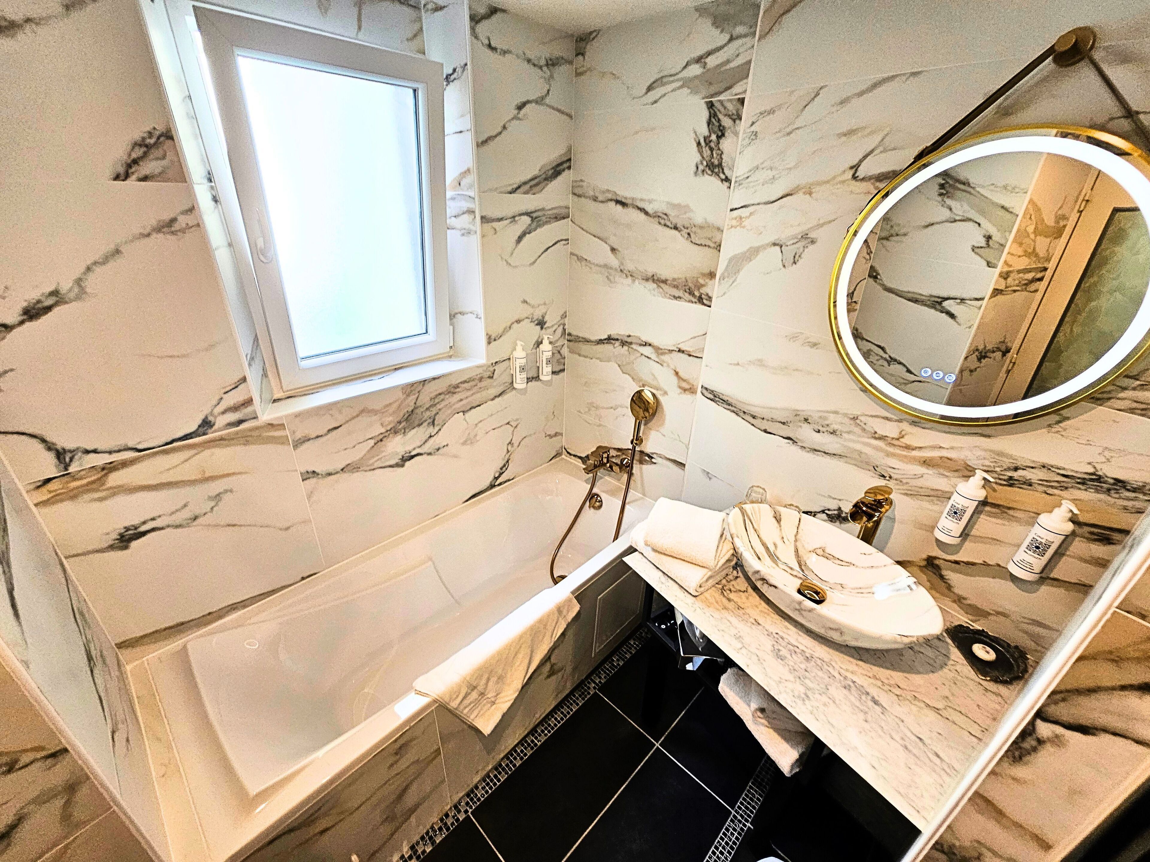 A bathroom with a marble wall, a bathtub, a sink with a marble countertop, and a mirror with a gold frame.