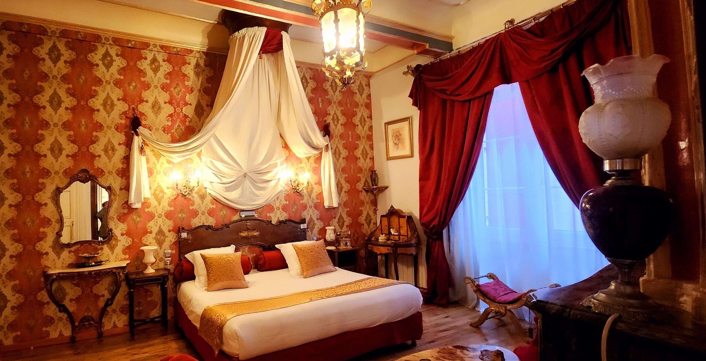 Una camera da letto con un letto a baldacchino, carta da parati decorata, un lampadario e un vaso di grandi dimensioni su un comodino.