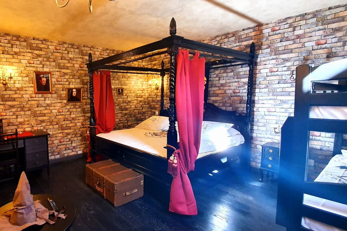 A four-poster bed with red curtains, a wooden chair, a small table with a bottle and a cloth, and a brick wall with a picture frame.
