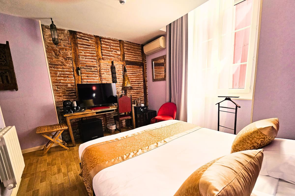 A hotel room with a brick wall, a bed with patterned bedding, a desk with a TV, and a red chair.