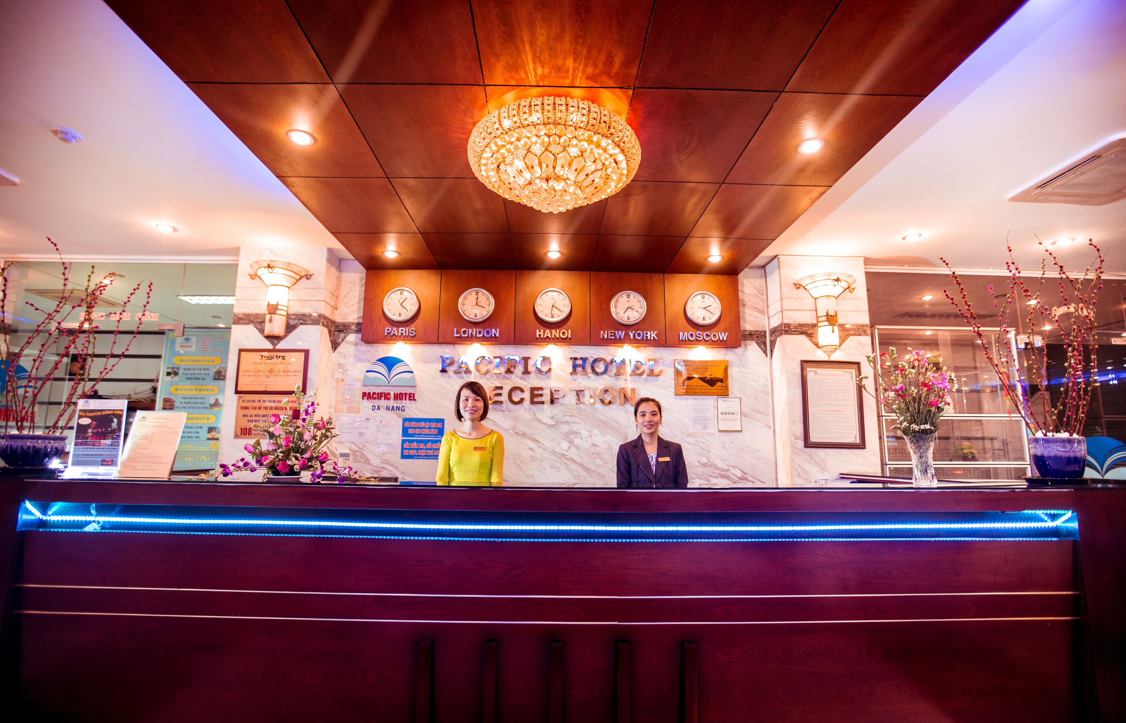 A reception desk at Pacific Hotel with a chandelier, clocks displaying different cities, and a reception area with a blue LED light strip.