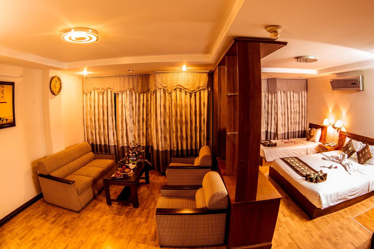 A hotel room with a sofa, two armchairs, a wooden coffee table, a bed with a nightstand, a lamp, and curtains.