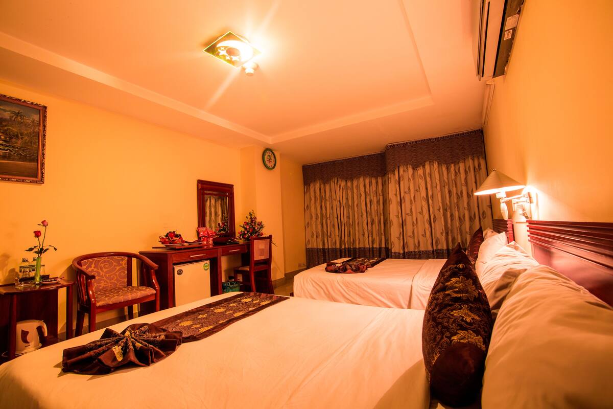 A hotel room with two beds, a desk, a chair, a clock, and a window with curtains.
