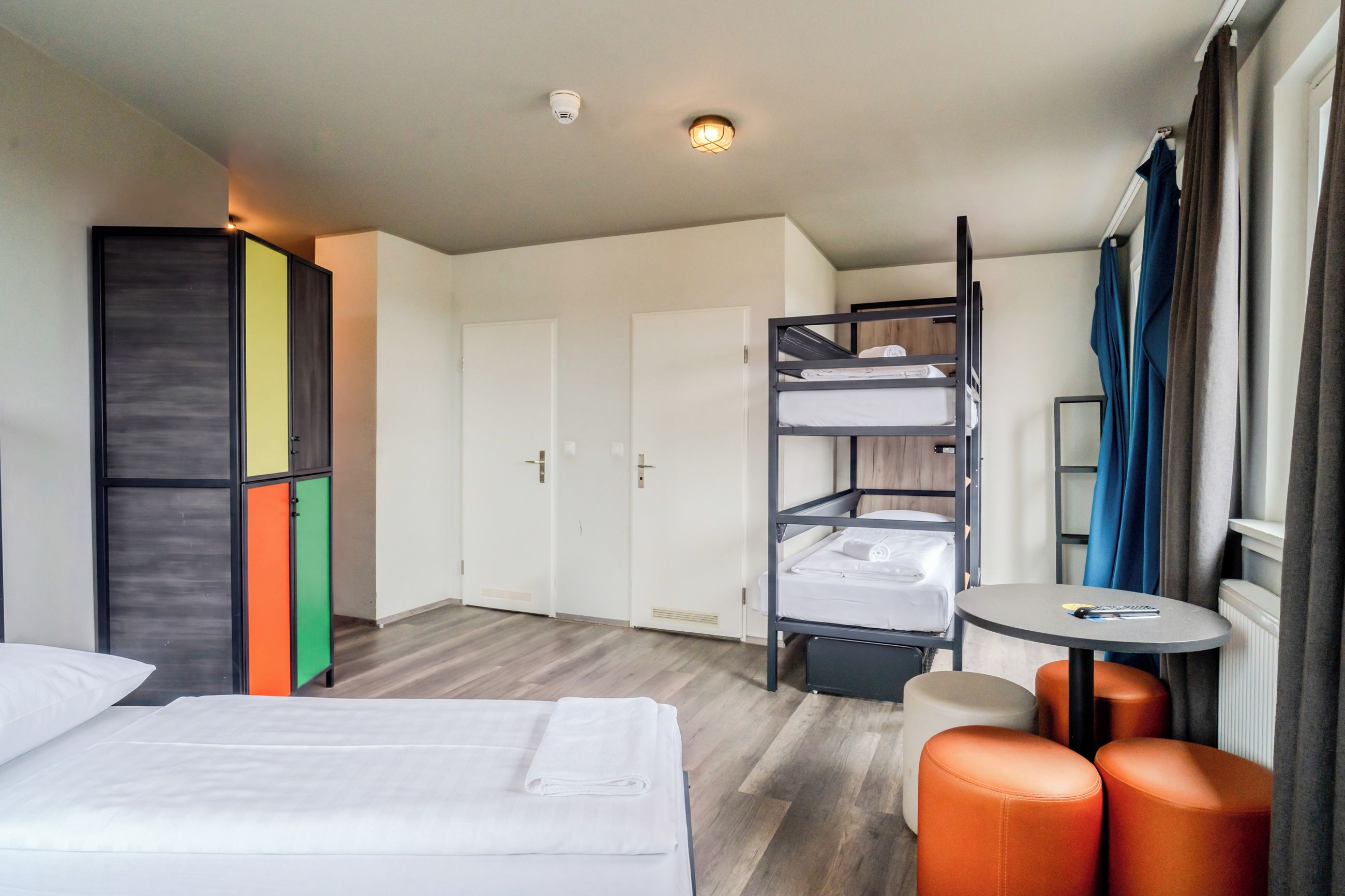 A hotel room with a bunk bed, a desk, a chair, a wardrobe, and a window with curtains.