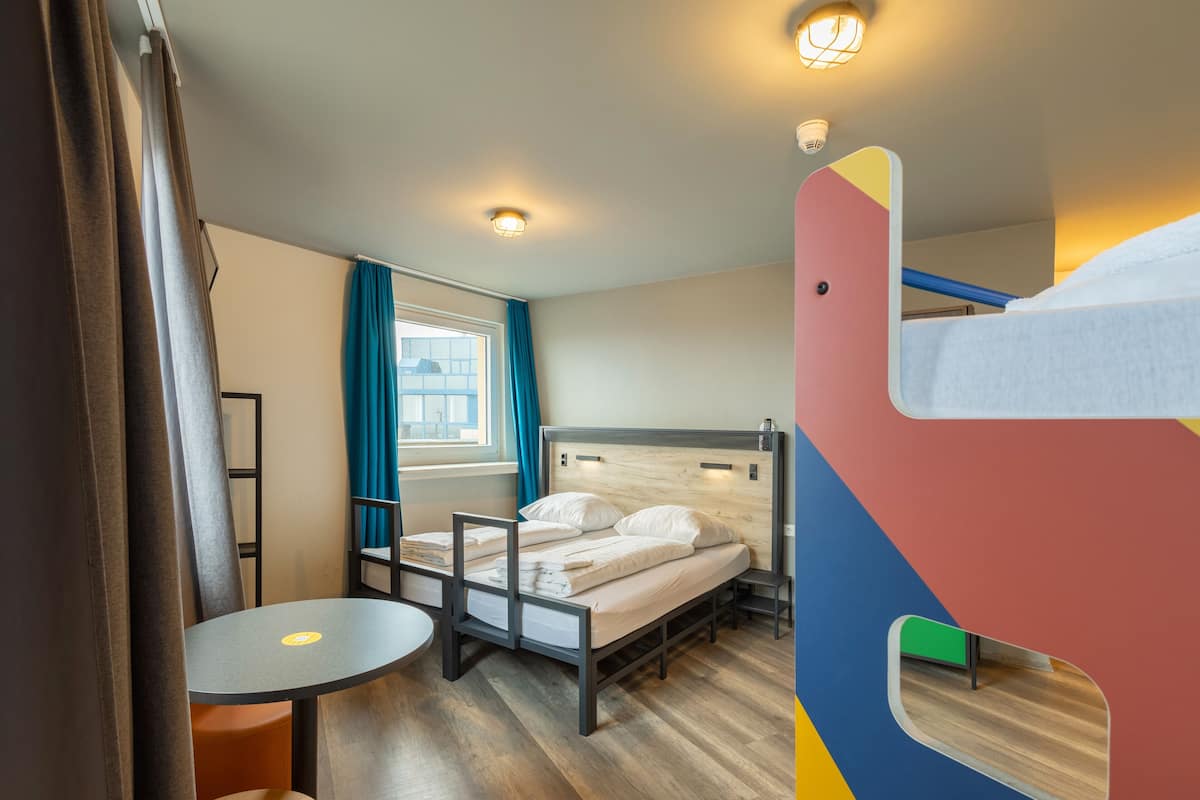 Une pièce comprenant un lit, une petite table, un lit superposé, une fenêtre avec des rideaux et un luminaire au plafond.