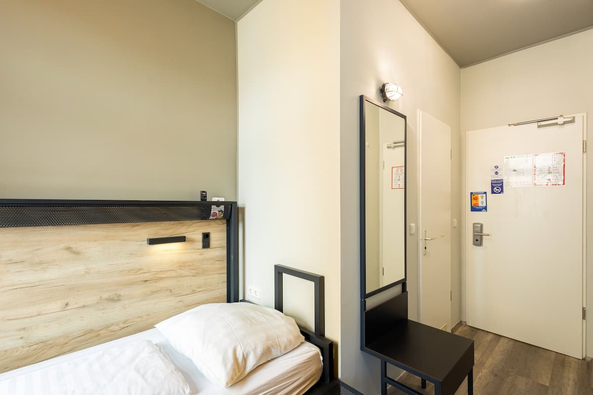 Une chambre à coucher avec une tête de lit en bois, un lit avec une literie blanche, une petite table noire, un miroir et une porte dotée d’une serrure et d’un avis.