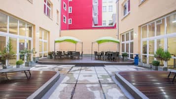 Un patio con mesas al aire libre, un escenario central y un edificio rojo al fondo.