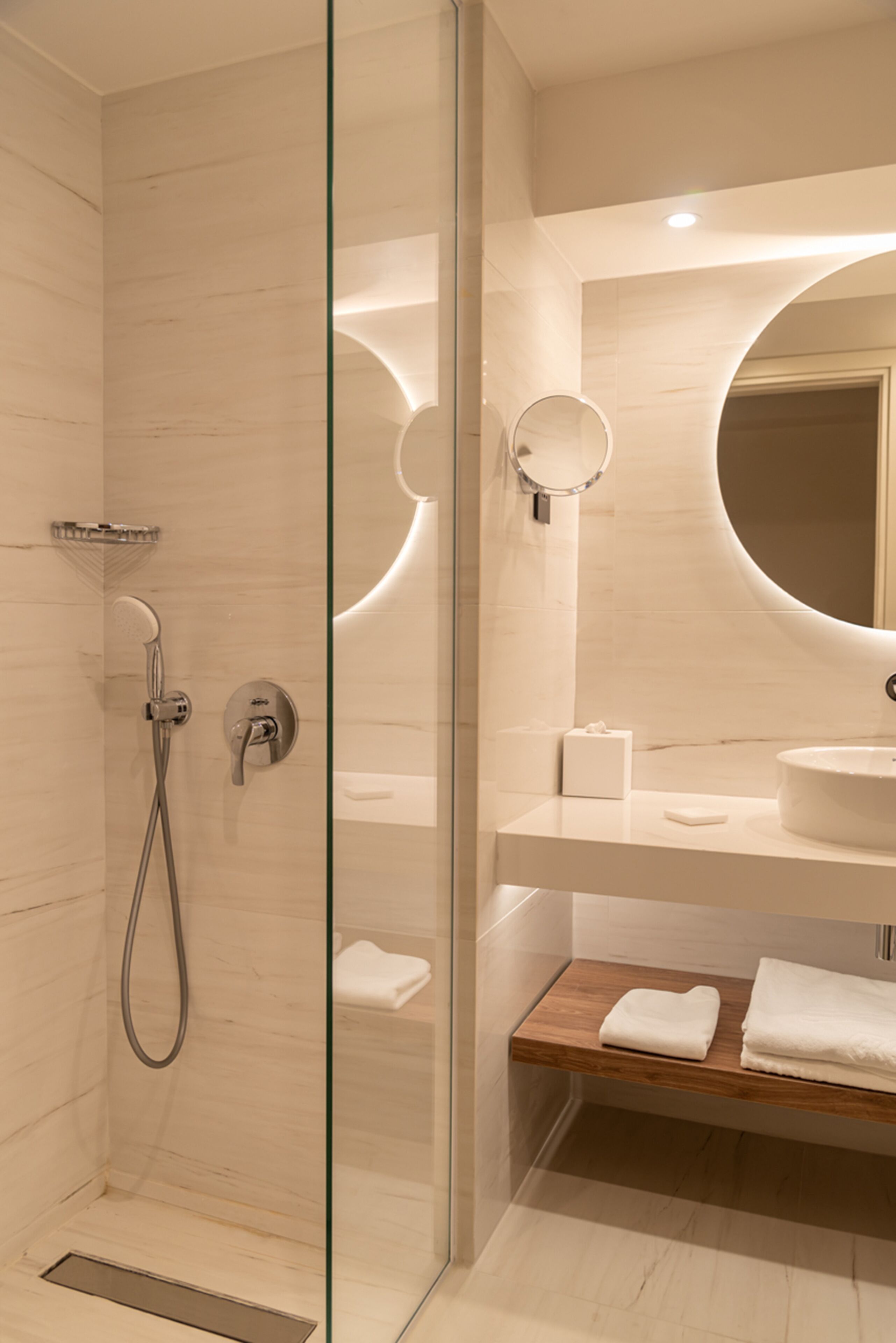 Une salle de bain moderne avec une douche à parois de verre, un miroir rond et une étagère en bois où sont posées des serviettes.