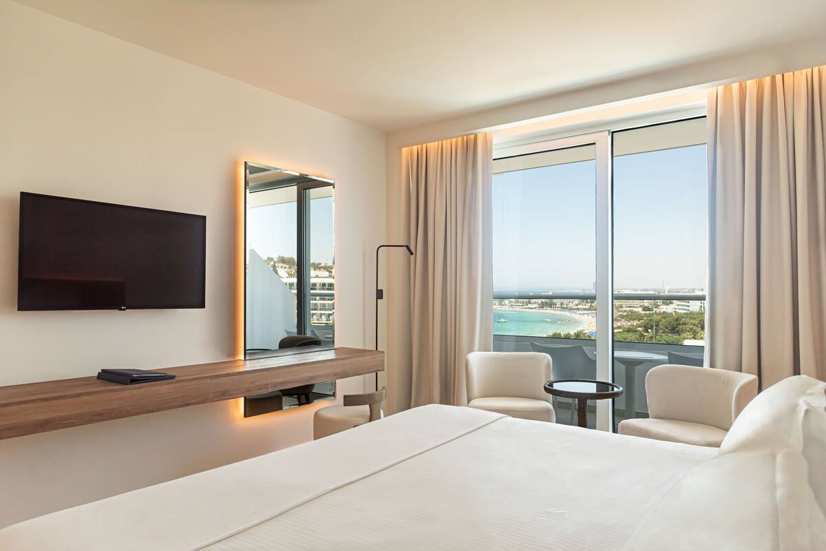 A hotel room with a large bed, a desk with a flat-screen TV, a chair, a small table, and a view of the beach and city through large windows.