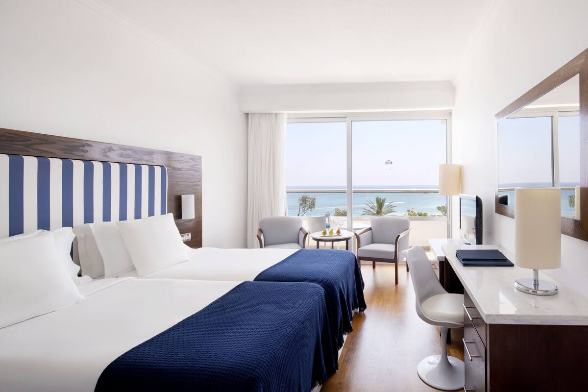 A hotel room with two beds, a desk, a chair, a small table with a lamp, and a view of the ocean.