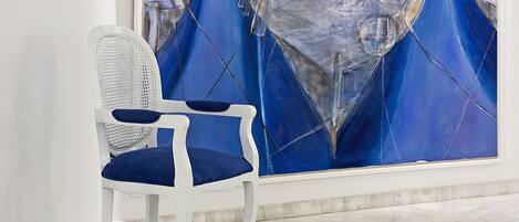 A room with a large abstract painting on the wall, a white chair with blue cushions, and a marble floor.