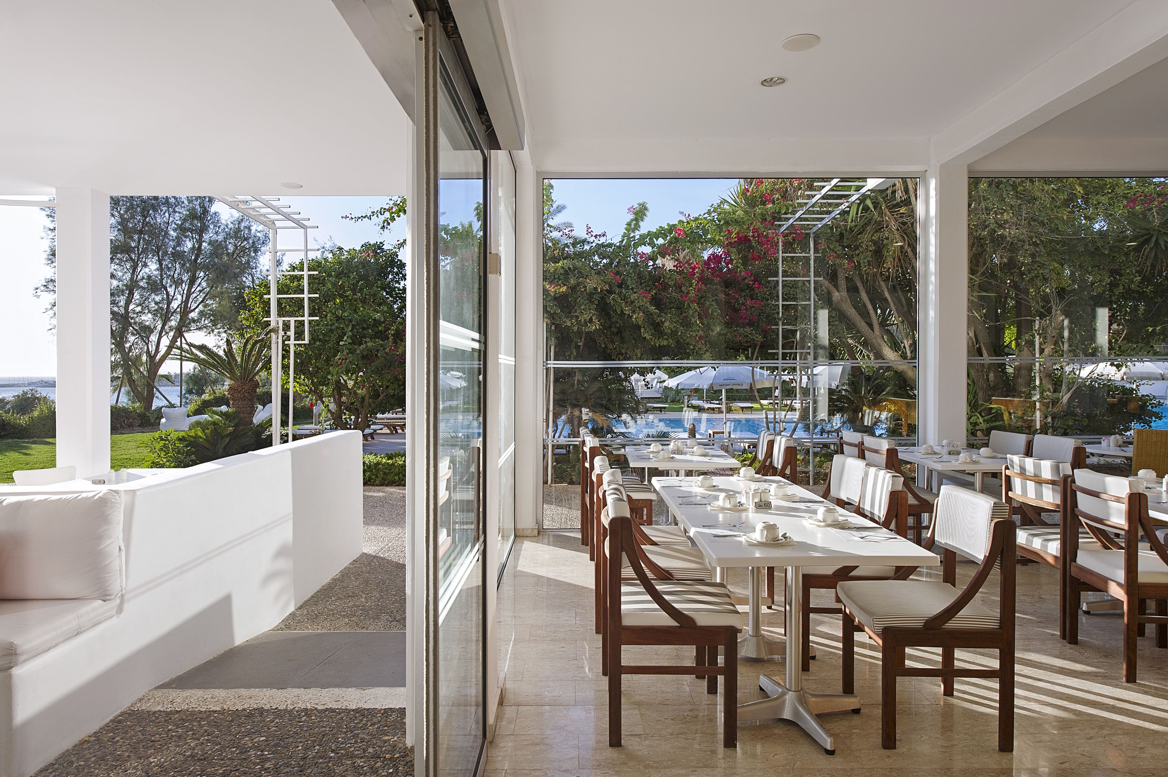 An outdoor dining area with tables and chairs, large windows, and a view of a pool and greenery.