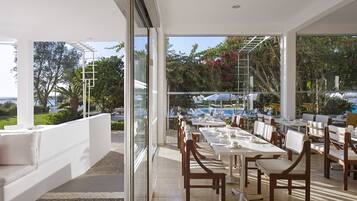 An outdoor dining area with tables and chairs, large windows, and a view of a pool and greenery.