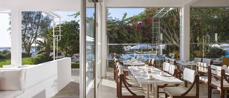 An outdoor dining area with tables and chairs, large windows, and a view of a pool and greenery.