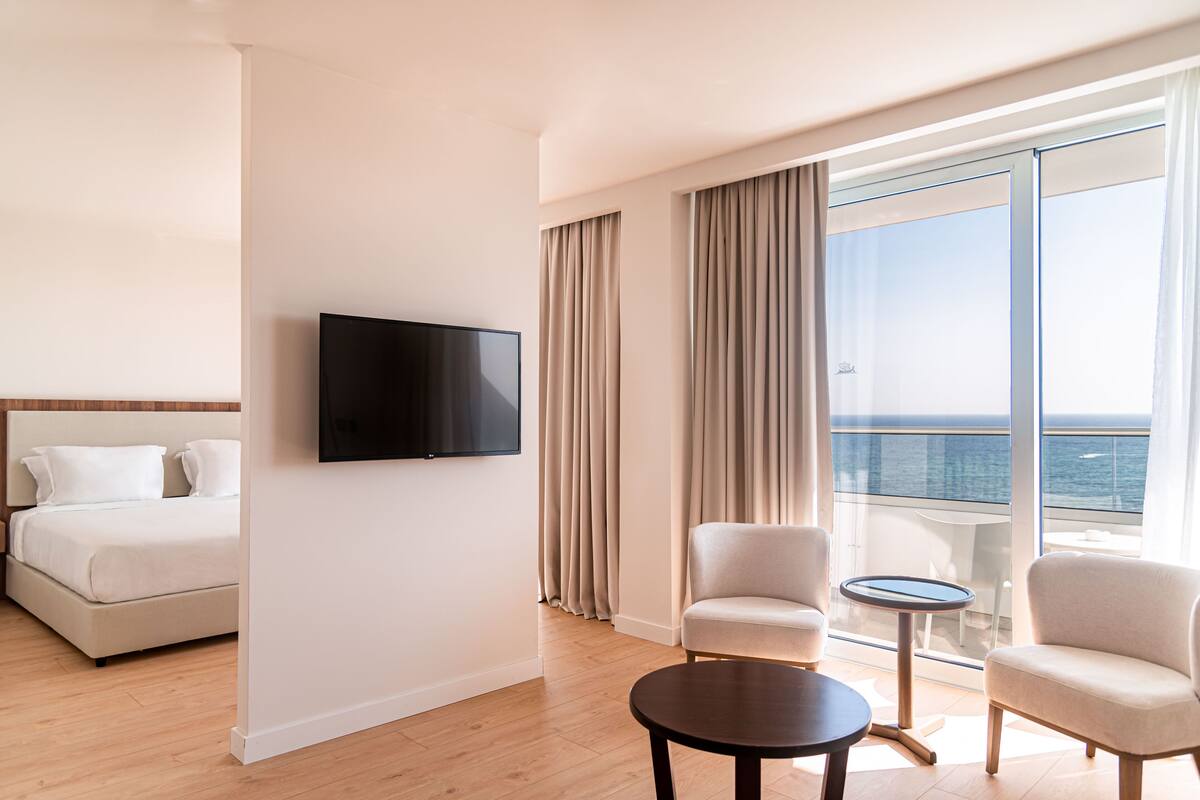 A hotel room with a bed, a TV, a balcony with a view of the sea, and two armchairs.