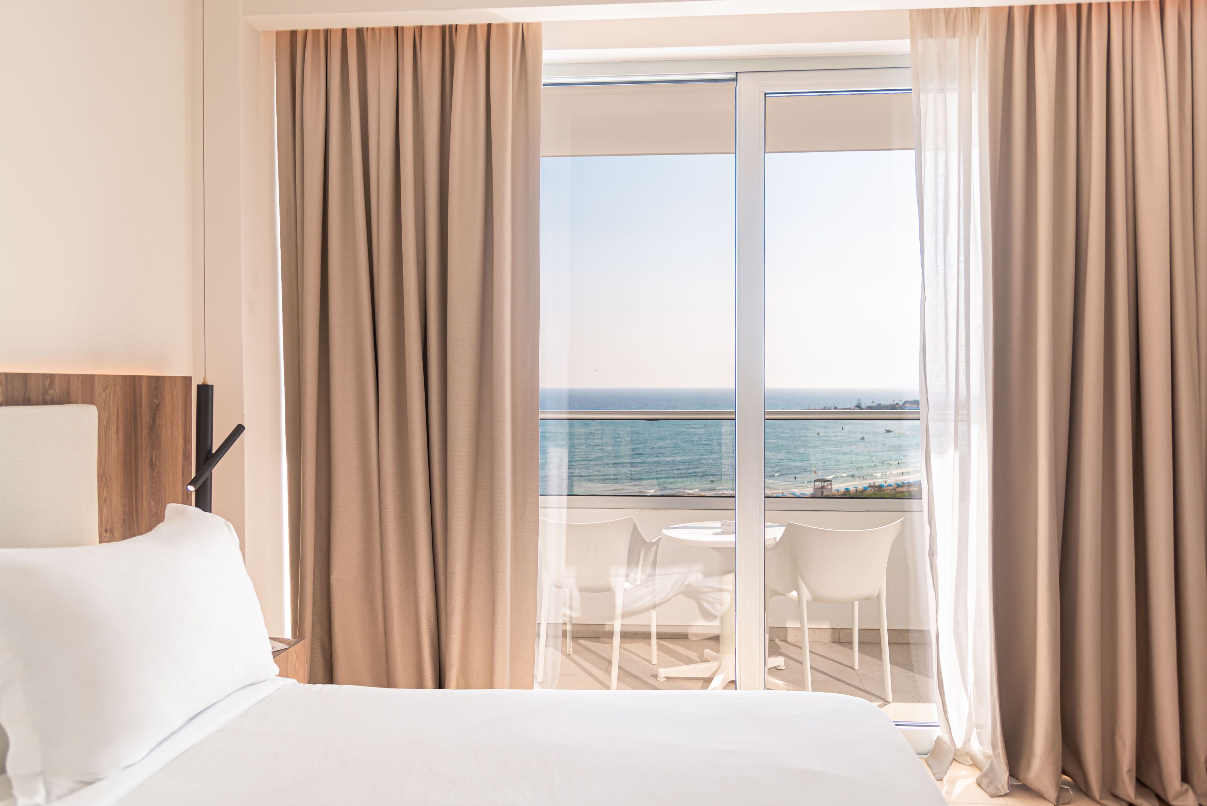 A hotel room with a bed, a window with curtains, and a view of the sea.