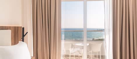 A hotel room with a bed, a window with curtains, and a view of the sea.