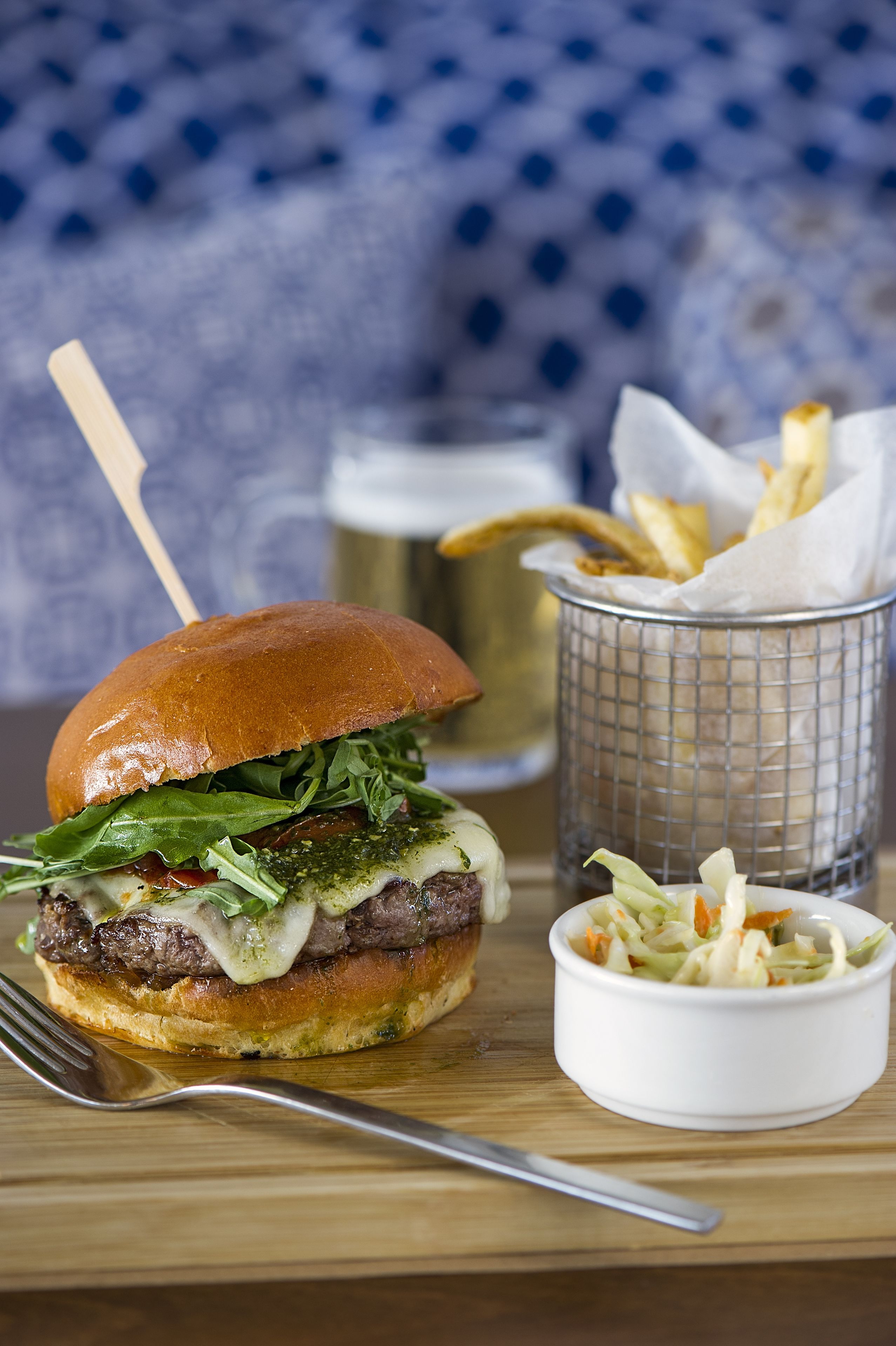 A burger with a bun, lettuce, cheese, and a toothpick. There is a basket of fries and a small bowl of coleslaw.