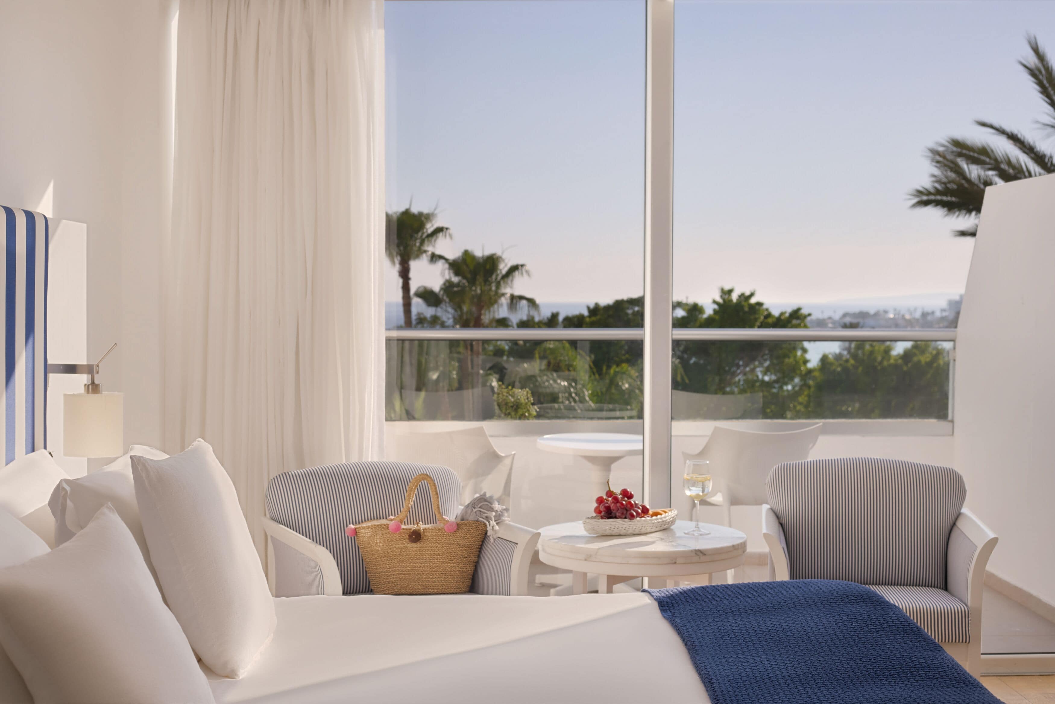 A modern hotel room with a balcony, white furniture, a table with a fruit bowl and glass, and a view of palm trees and the ocean.